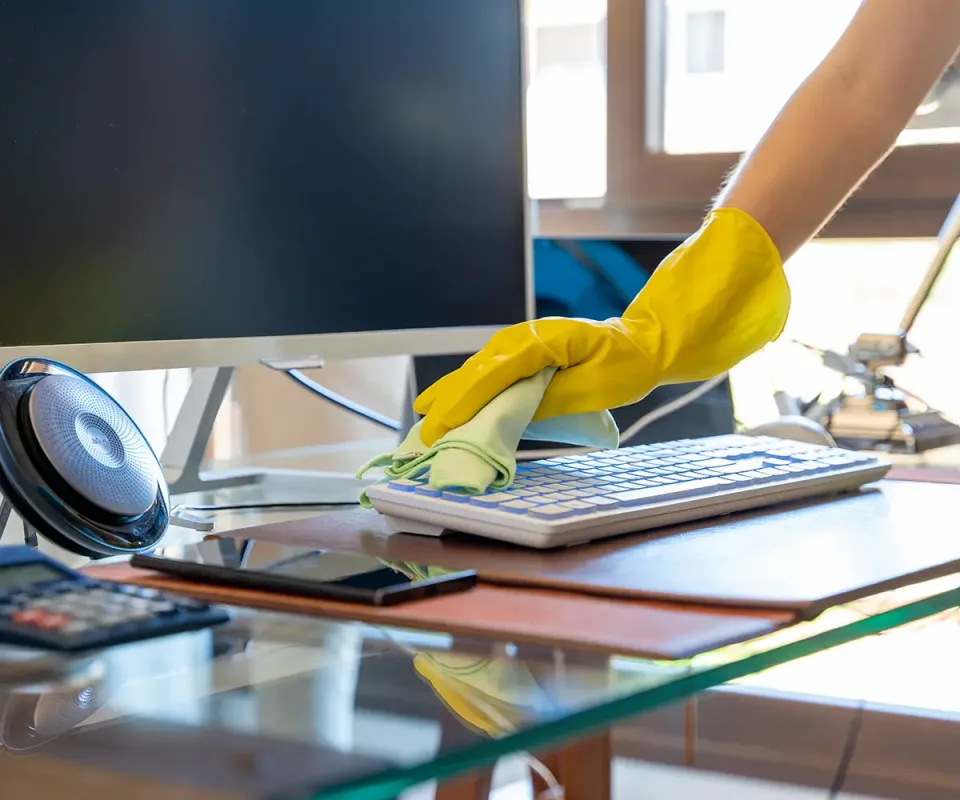 A gloved hand wiping a keyboard on an office desk beside a monitor