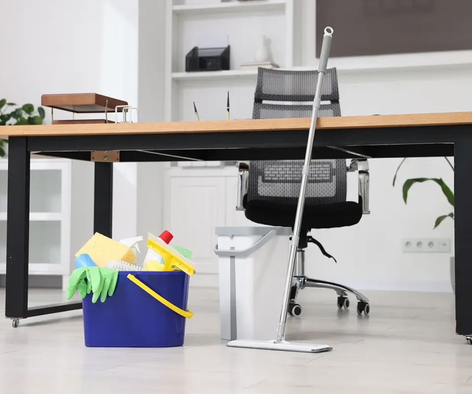A mop, a wringer bucket and a caddy of cleaning supplies beside a desk and an office chair